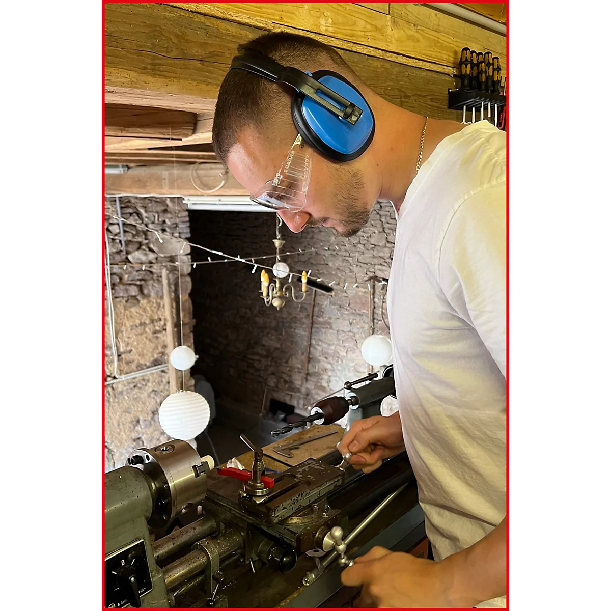 Mann mit Gehörschutz und Schutzbrille an der Drehbank. Er arbeitet an Metall, der Hintergrund zeigt einen Steinbogen.